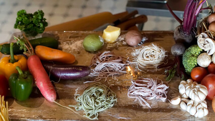 A wooden cutting board displays an assortment of fresh vegetables and colored pasta