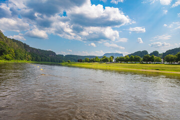 Scenic summer view of the Elbe River at Rathen, Saxon Switzerland. near the Bastei. Traditional houses, forested sandstone cliffs, and low water levels define this peaceful German river landscape.