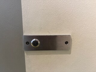 Minimal close-up of a metallic push button mounted on a textured white wall, secured with screws. 