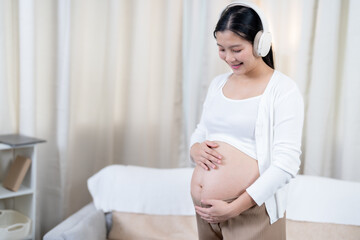 Asian pregnant woman wearing white sweater brown pants smiling listening to music with headphones...
