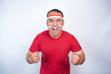 Fototapeta premium A young Asian man with a happy successful expression, raised a fist wearing red shirt and flag headband isolated by white background. Indonesia's independence day concept.