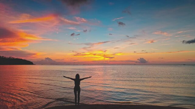Silhouette of a young woman standing on a beach with open arms, embracing the vibrant and colorful sunset over the sea in Thailand, captured through stunning aerial drone footage