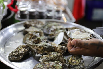 Close-up of a tattooed vendor's hands skillfully opening fresh oysters with a knife at a seafood...