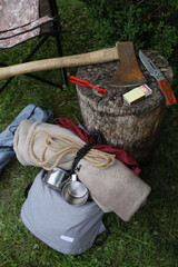 Hiking. Things and objects for tourism and survival in the forest or mountains. On the grass near the stump lie an axe, matches, canned food, rope, a tourist chair, a backpack and other things