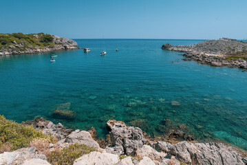 A calm bay with turquoise water and anchored boats surrounded by rocky cliffs and sparse vegetation on the island of Rhodes in Greece.