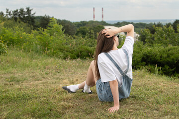 A young girl in a white shirt and denim overalls sits on grass, looking out at a distant landscape featuring factory smokestacks. The scene captures a moment of relaxation in nature