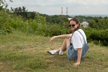 A young woman sits comfortably on the grass, wearing denim overalls and sunglasses. She smiles at the camera with a beautiful green landscape and industrial smokestacks in the background