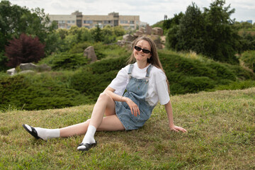 A young woman wearing denim overalls and sunglasses sits comfortably on a grassy hill in an urban park. The backdrop features a modern building and lush greenery, suggesting a serene atmosphere