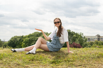 A young woman dressed in a white shirt and denim overall sits comfortably on a grassy knoll, smiling and enjoying a sunny day. Trees and a building are in the background