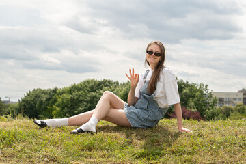 A young woman sits on a grassy hill, waving and smiling. She wears sunglasses and casual summer attire, enjoying a relaxed afternoon under a partly cloudy sky in an urban park