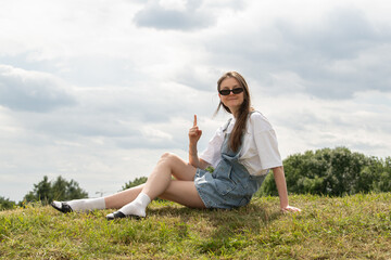 A young woman sits comfortably on a grassy slope, playfully pointing while wearing a casual outfit of a white shirt and denim overalls. The cloudy sky adds a relaxed vibe to the summer day