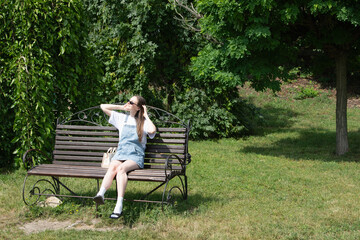 A young woman relaxes on a park bench, soaking up the sun while dressed in a casual outfit with sunglasses and denim. Lush greenery surrounds her, creating a serene atmosphere