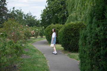 A young woman strolls along a winding path in a rose garden, surrounded by lush greenery and blooming flowers under a clear sky. The scene exudes a calm, serene atmosphere