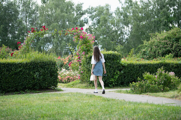 A woman strolls through a colorful flower garden filled with blooming roses and lush greenery on a sunny summer day. The pathway leads her into the picturesque scene