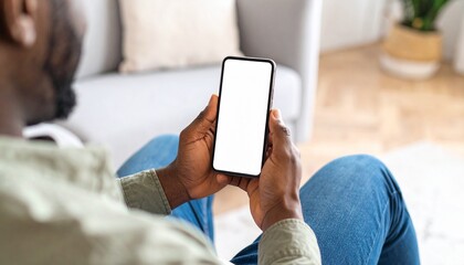 African American man holding a smartphone with a mockup white blank display, empty screen for app ads sitting on a couch at home. Mobile applications technology concept, over-the-shoulder close-up.