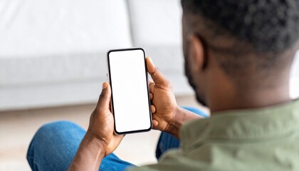 African American man holding a smartphone with a mockup white blank display, empty screen for app ads sitting on a couch at home. Mobile applications technology concept, over-the-shoulder close-up.