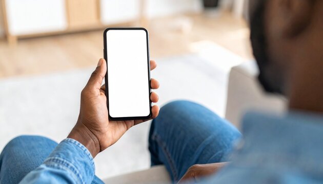 African American man holding a smartphone with a mockup white blank display, empty screen for app ads sitting on a couch at home. Mobile applications technology concept, over-the-shoulder close-up.