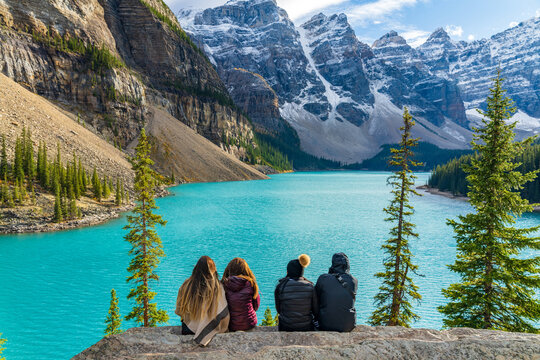 Four young women on vacation, sitting together by the turquoise Moraine Lake, admiring the snow-capped Ten Peaks. Banff National Park, Alberta, Canada.