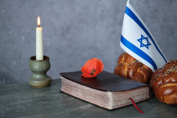 Israeli flag with an a closed Koran decorated with poppy flower, traditional challah bread, candle on a gray background. highlighting a cultural and religious gathering. Jewish Remembrance Days. 