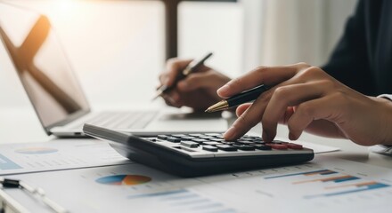 Person calculating data on a calculator while holding a pen, with laptop and charts in the background.