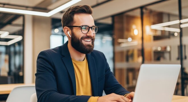 Man wearing glasses and earbuds smiles while working on a laptop in an office
