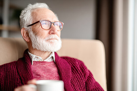 An elderly man with white hair and beard, wearing glasses and a red sweater, sits comfortably holding a mug, gazing thoughtfully out of a window.
