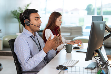 Professional customer support representative wearing headset is engaged in conversation while working at desk with computer. colleague is seated nearby, focused on her tasks