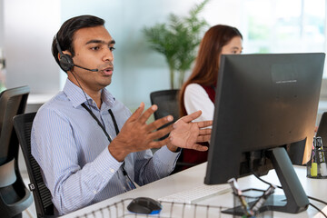 Customer support representative wearing headset is actively engaging in conversation while working on computer in modern office environment. colleague is visible in background