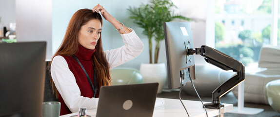 Focused woman working in modern office environment, using computer and laptop, possibly engaged in tech related customer support tasks. setting includes desk, monitor arm, and indoor plants