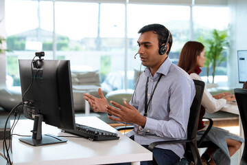 Professional tech support representative wearing headset is assisting customer remotely while working at modern office desk. background shows collaborative workspace with another employee
