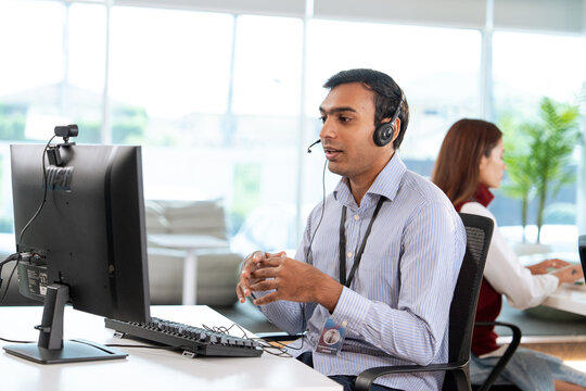 Professional tech support representative wearing headset is assisting customer at modern office desk, with computer and keyboard in front. colleague is working in background
