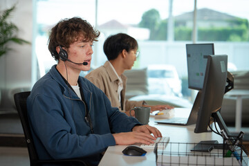 Two young professionals are working modern office environment. foreground shows man wearing headset, focused providing tech customer support while typing keyboard. In background, another