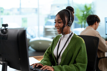 A cheerful customer support representative wearing headset and green sweater is working at computer in modern office. colleague is visible in background, CRM collaborative atmosphere