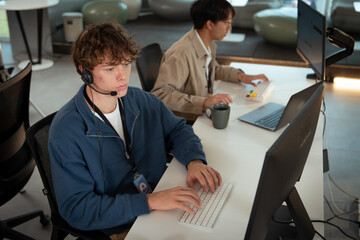 Two young professionals working modern office environment, focusing customer support tasks. One is wearing headset, typing keyboard, and engaging tech related assistance, while other works laptop