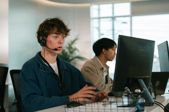 Young man wearing headset provides tech customer support at modern office, working attentively on computer alongside colleague. environment is bright and professional