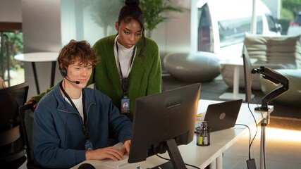 Two customer support professionals are working together modern office environment. One is seated, wearing headset, and typing computer, while other stands nearby, attentively observing screen