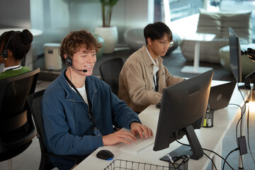 A two young professionals are working modern office environment, providing tech customer support. one is wearing headset and typing keyboard, while other is focused his computer. atmosphere