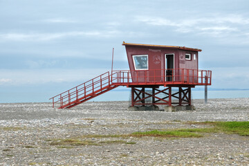 Red lifeguard tower on stilts overlooks a rocky beach and calm sea under a moody, cloud-filled sky.