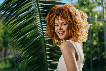 smiling woman with curly hair outdoors wearing white tank top near tropical palm leaves in sunny...