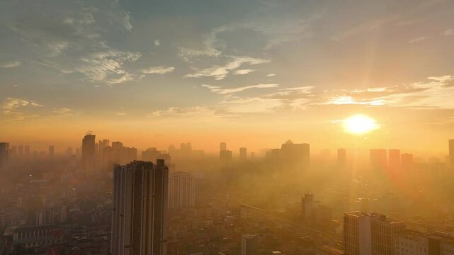 An atmospheric aerial view of Hanoi, Vietnam, at sunset, bathed in the warm glow of the sun through a soft haze. The sprawling urban landscape stretches towards the horizon, with distant lakes or rive