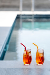 Two homemade Aperol spritz cocktails with ice and colorful silicone straws sit on a sunny poolside...