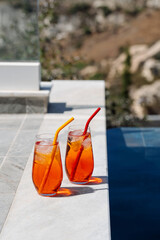Two homemade Aperol spritz cocktails with ice and colorful silicone straws sit on a sunny poolside...