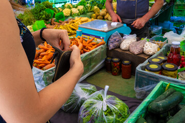 Customer purchasing fresh vegetables at outdoor market stall with display of carrots, cucumbers,...