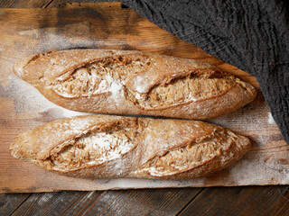 Two loaves of rustic bread placed on a wooden surface, with a knitted cloth partially covering the...