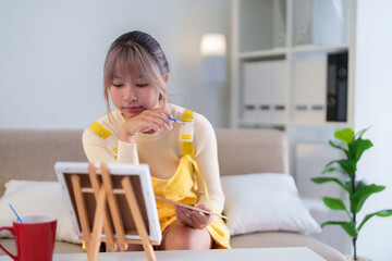 Young Asian woman engaged in painting her artwork on canvas with brush to express creativity and...