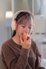 Young Asian woman wearing headphones enjoys eating a fresh apple indoor.