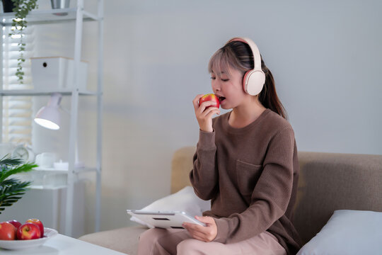 Asian woman enjoys an apple while relaxing with tablet at home wearing headphones - Powered by Adobe