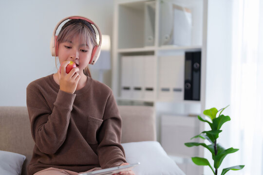Young Asian woman eating an apple while listening with headphones Indoor relaxing scene - Powered by Adobe
