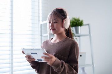 Fototapeta premium Young Asian woman enjoys tablet with headphones at home