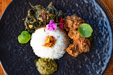 Overhead shot of a black plate with rice, meat, vegetables, and sauce, garnished with flowers and...
